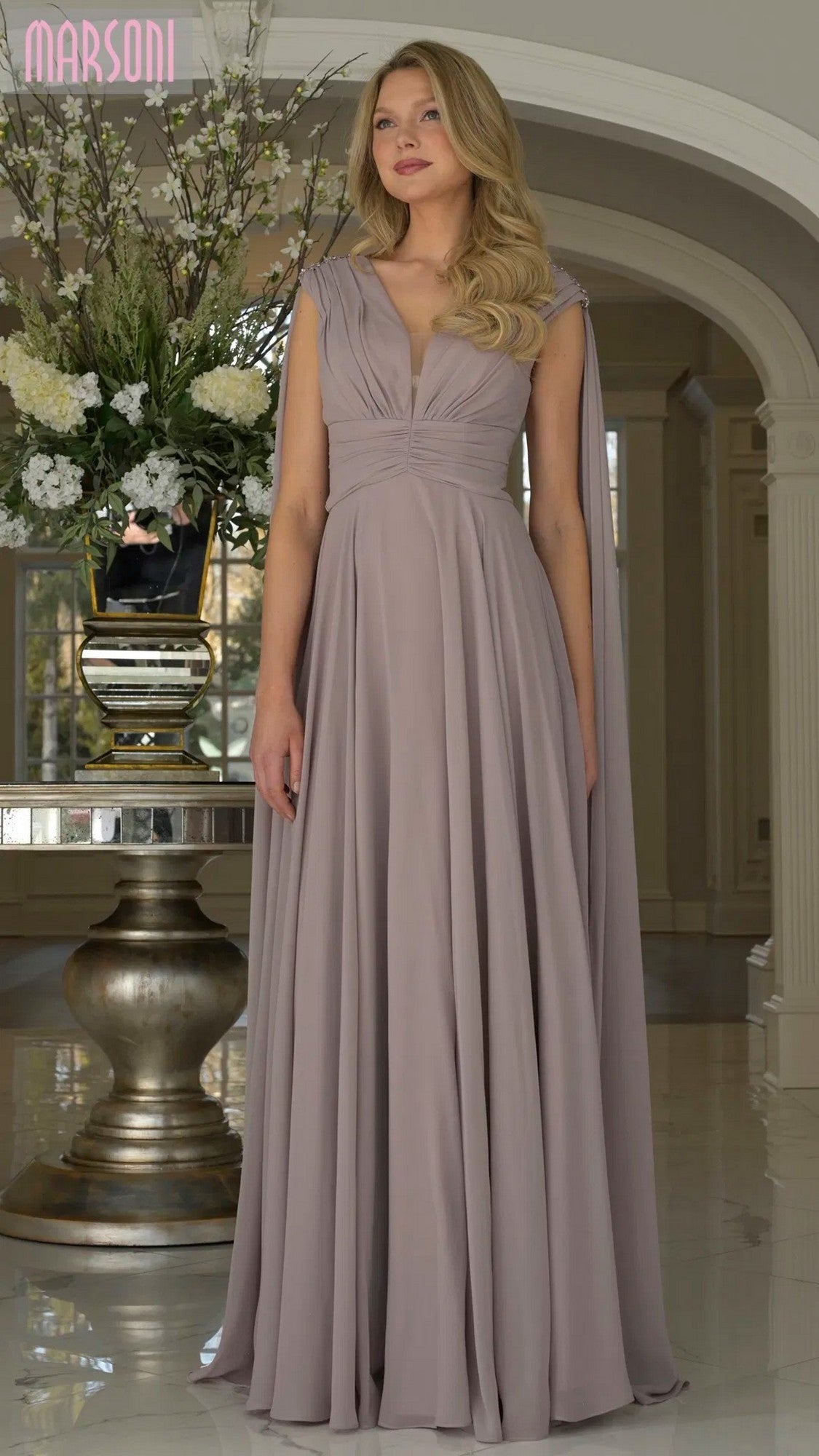 A woman stands indoors wearing the Marsoni MV1349 long formal dress in dusty rose, featuring chiffon fabric, long cape sleeves, draped shoulders, and a floor-length design. She poses before a table with a large floral arrangement.